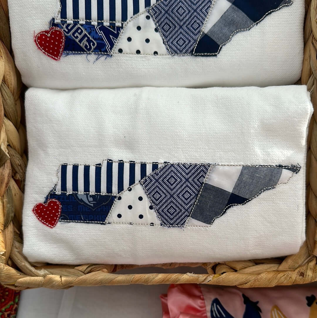 100% cotton tea towel featuring an appliqué of the state of tennessee in shades of blue fabric with a red heart sewn where memphis is - in the bottom left corner of the state.